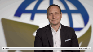 A man in a dark suit sits and smiles in front of a blurred background featuring a blue and gold globe design, with a video player bar visible at the bottom of the image.